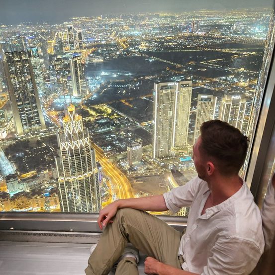 Ein Mann sitzt in einer verglasten Aussichtsplattform und betrachtet bei Nacht eine Stadtsilhouette. Hohe Gebäude und Straßen mit beleuchteten Autos umgeben ihn.