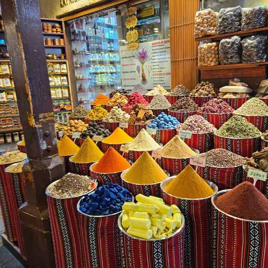 Ein Gewürzladen mit verschiedenen farbenfrohen Gewürzen in Eimern, darunter Safran, Paprika und Kurkuma. Im Hintergrund befinden sich Regale mit verpackten Waren.