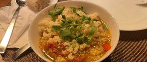 Eine Schüssel Couscous-Suppe mit frischen Kräutern als Garnierung steht auf einem gewebten Tischset, neben einem Löffel und einem Teller.