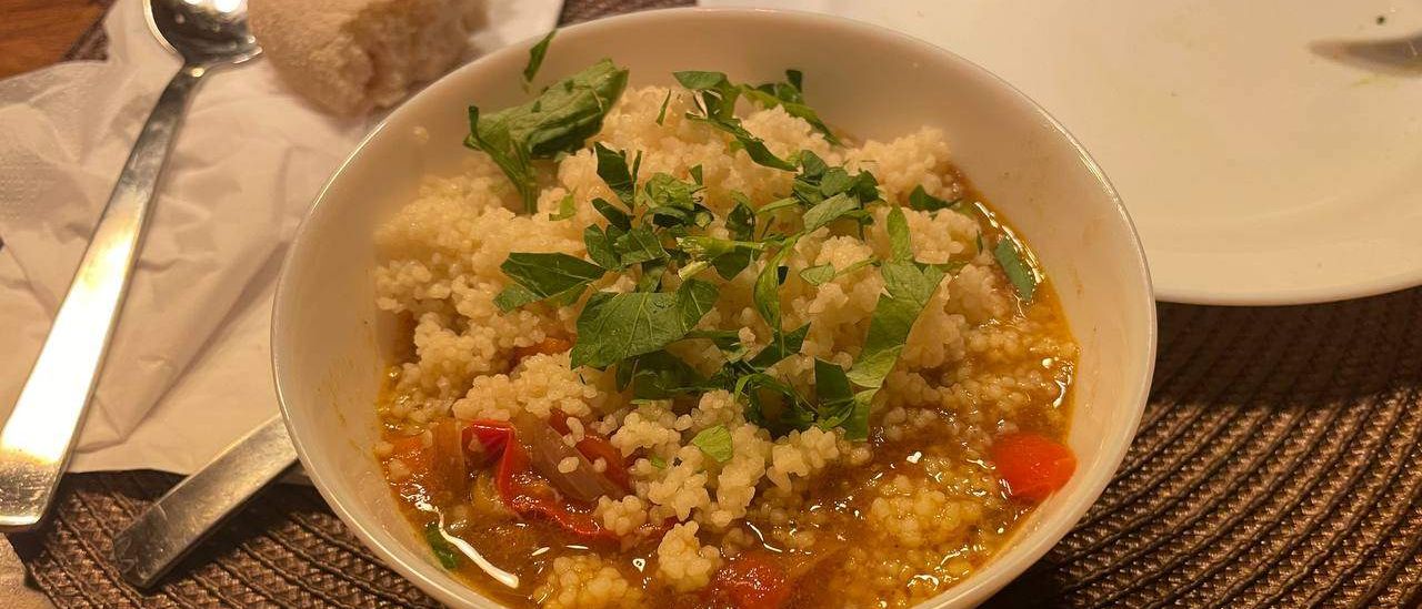 Eine Schüssel Couscous-Suppe mit frischen Kräutern als Garnierung steht auf einem gewebten Tischset, neben einem Löffel und einem Teller.