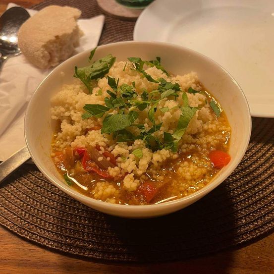 Eine Schüssel Couscous mit Gemüse, übersät mit Kräutern, steht auf einem gewebten Platzset auf einem Holztisch. Ein Löffel und eine Serviette sind in der Nähe.