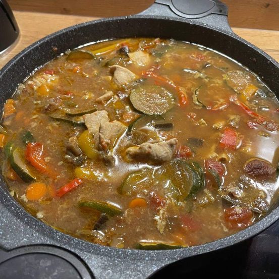Eine Gusseisenpfanne auf einem Herd hält einen reichhaltigen Eintopf mit Fleischstücken, Zucchini, Paprika und Tomaten.