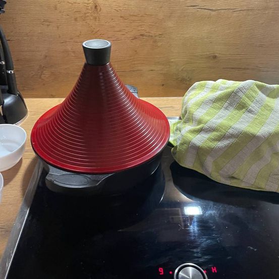 Ein roter Keramiktopf mit konischem Deckel steht auf einem schwarzen Induktionsherd. Ein grün-weiß kariertes Handtuch liegt daneben. Links steht eine weiße Schüssel.