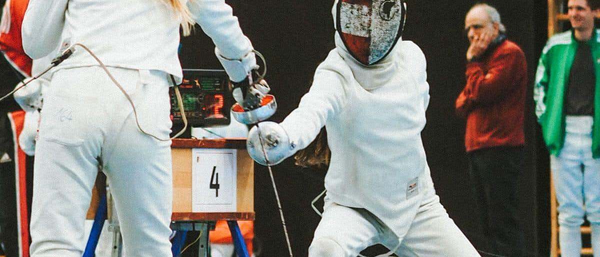 Zwei Fechterinnen kämpfen, eine mit der Nummer '4'. Zuschauer schauen zu, einer trägt einen roten Mantel. Der Platz ist mit einem Basketballkorb ausgestattet.