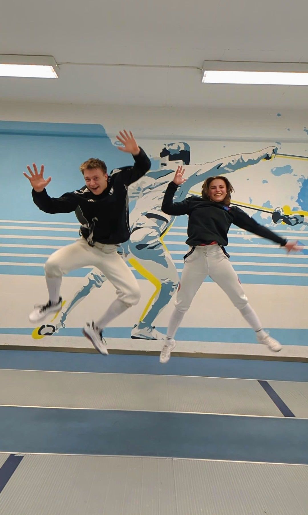 Two people jump in the air with smiles, wearing fencing uniforms, with a fencing mural in the background.
