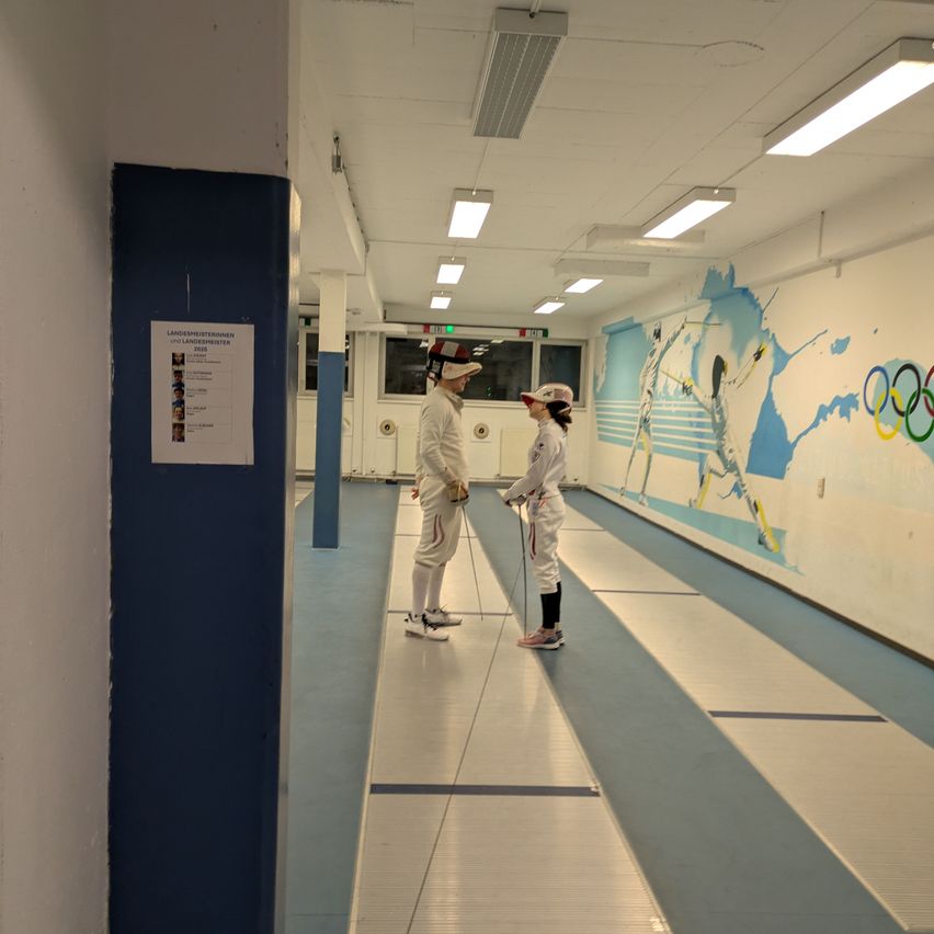 Two people in fencing uniforms face each other in a corridor. The walls are painted with blue and white Olympic themes.