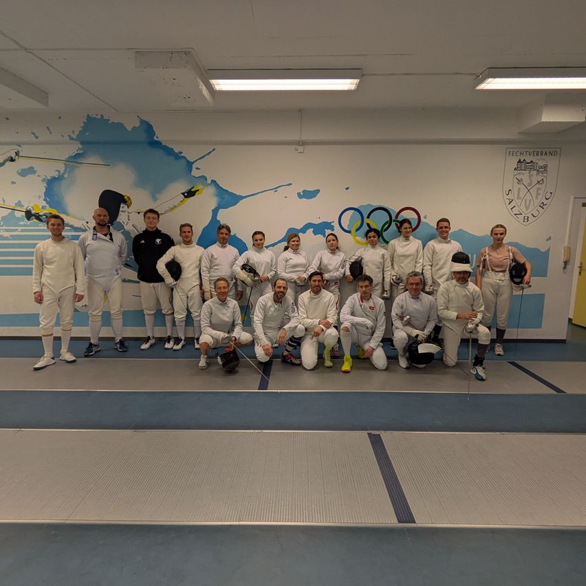 A fencing team poses for a photo in a room with a mural of skiers and Olympic rings on the wall.