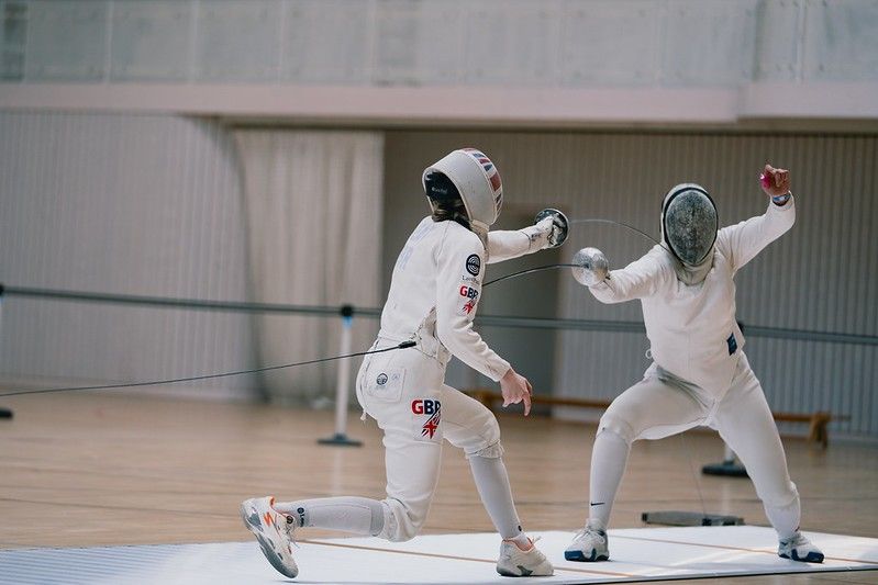 Zwei Fechter in weißen Uniformen mit GBR-Patches führen einen Fechtkampf auf einem Holzboden aus. Ein Fechter ist bereit zuzustechen.
