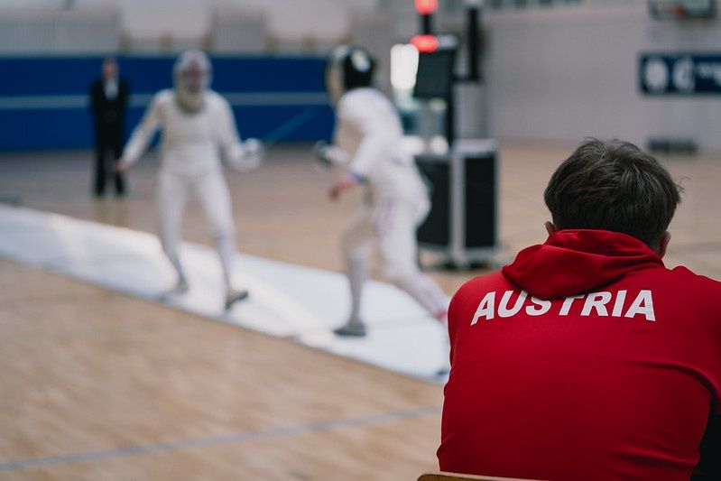 Zwei Fechtathlethen duellieren sich in einem Wettkampf, während ein Mann in einem roten Trikot mit dem Aufdruck 'AUSTRIA' von der Seitenlinie zusieht.