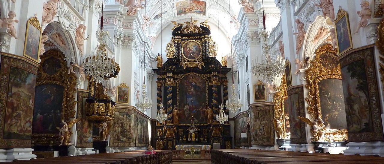 Ein prachtvoller Kircheninnenraum mit verzierten hölzernen Kirchenbänken, goldenen Altären und kunstvollen Fresken und Skulpturen schmücken Wände und Decke.