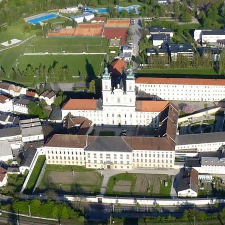 Luftaufnahme eines großen, historischen Gebäudekomplexes umgeben von Gärten, Tennisplätzen und einem Sportplatz. Der Komplex umfasst mehrere Gebäude, einen großen Innenhof und eine nahegelegene Eisenbahnstrecke.
