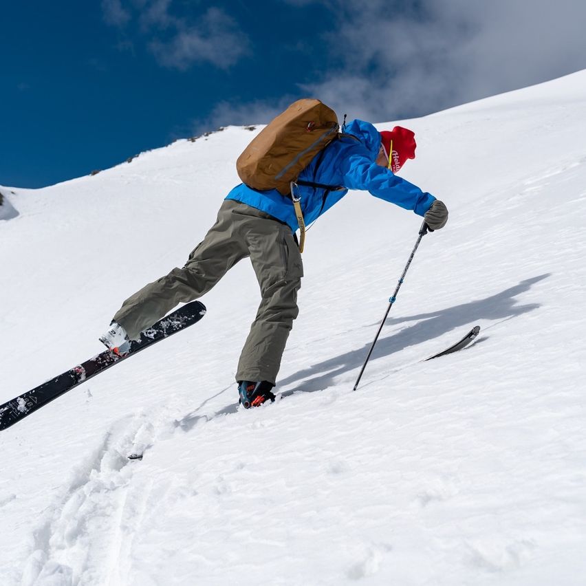 Ein Skifahrer mit Rucksack fährt einen schneebedeckten Berghang hinunter, trägt einen blauen Mantel, eine rote Mütze und Handschuhe und hat Skistöcke in der Hand.