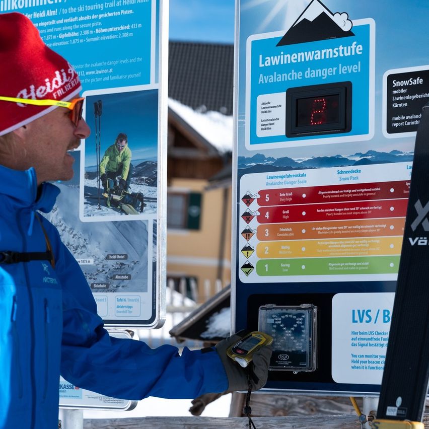 Ein Mann in einer blauen Jacke und einem roten Hut untersucht ein Lawinenwarntafel und benutzt ein Handgerät. Die Tafel zeigt Stufen von 1 bis 5 und eine digitale Anzeige.