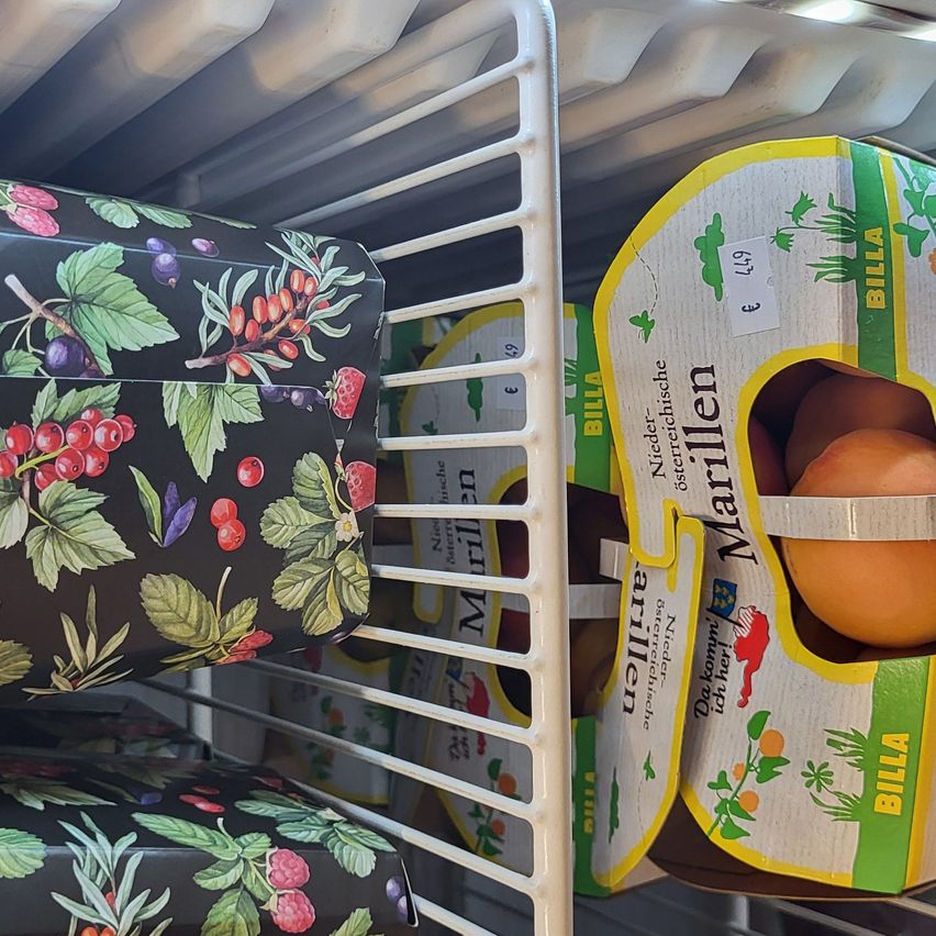 Obstkartons im Kühlschrank, darunter ein Kasten mit Erdbeeren und ein Kasten mit Orangen, mit Etiketten, die Preis und Herkunft angeben.