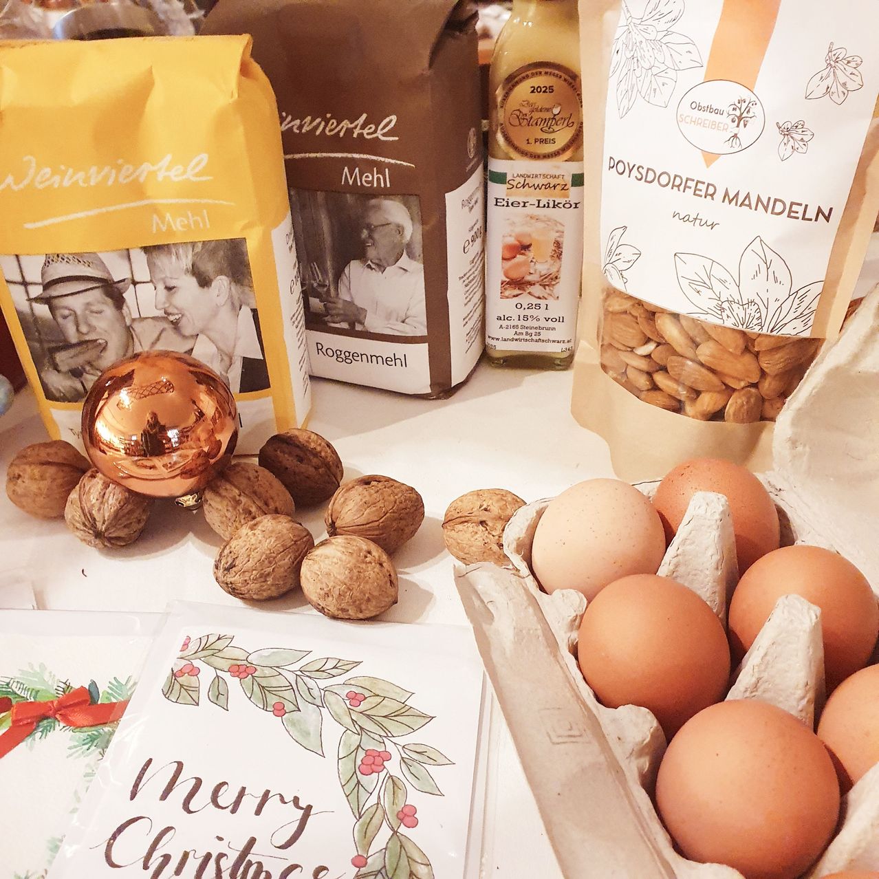 Auf einem Tisch sind verschiedene Feiertagsartikel zu sehen: eine Kupferkugel, Walnüsse, Eier, ein Mehl-Mehlsack, eine Flasche Eier-Likör und ein Beutel Poysdorfer Mandeln.