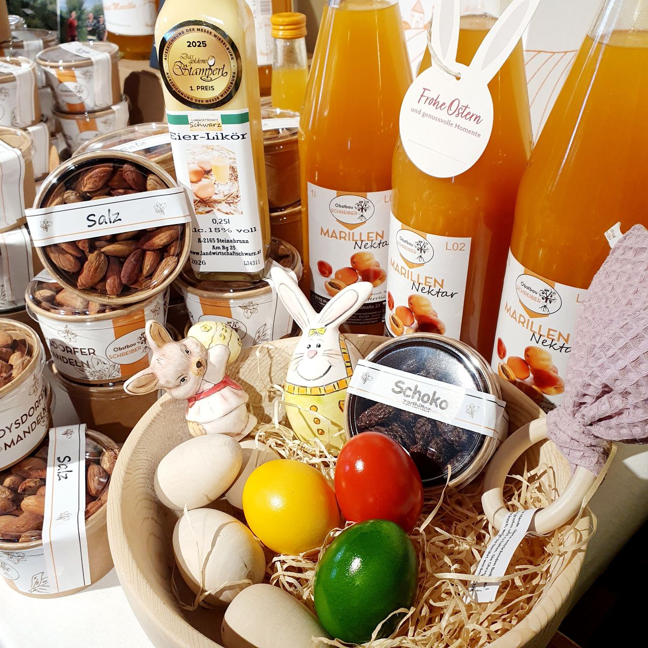 Eine Holzschale mit Ostereiern, Nüssen und getrockneten Früchten. Verschiedene Flaschen Nektar, darunter Aprikose, und Gläser mit Honig. Dekorative Osterhasen und andere Gegenstände.
