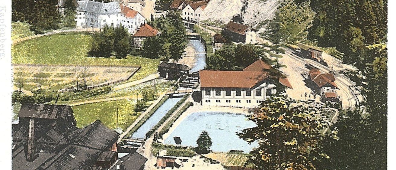 Ein altes Postkartenbild zeigt ein malerisches Dorf in den Bergen, mit einem Fluss, der hindurchfließt und einem großen Gebäude am Wasser.