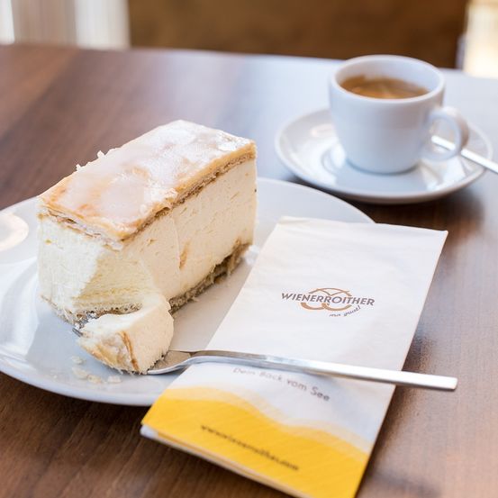 Ein Stück Käsekuchen auf einem Teller mit einer Gabel, einer Serviette und einer Tasse Kaffee auf einer Untertasse auf einem Holztisch.