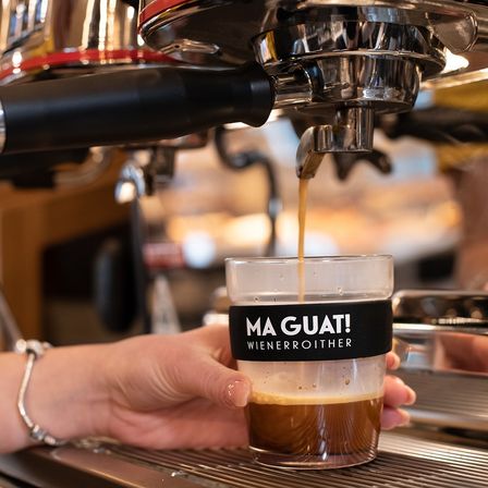 Eine Person hält eine Tasse mit der Aufschrift MA GUAT! WIENERROITHER, in die Kaffee aus einer Maschine gegossen wird.