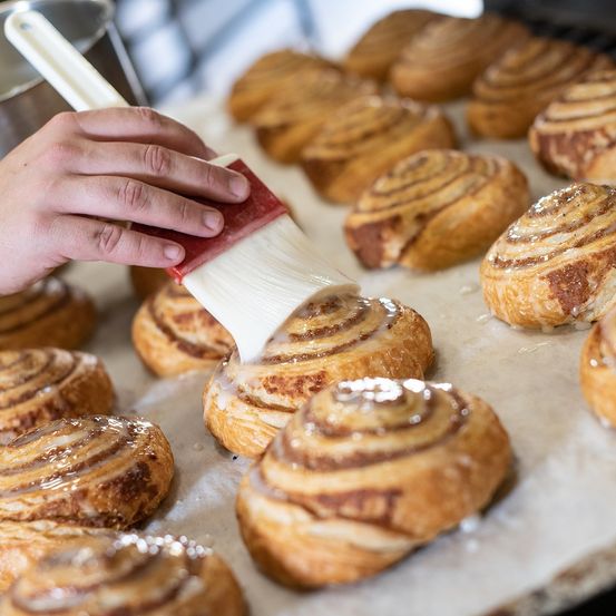 Eine Hand glazuriert goldene Zimtschnecken mit Zuckerguss auf einem Backblech. Verschiedene andere Brötchen sind verstreut.