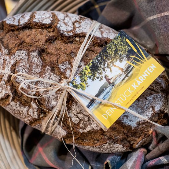 Ein Laib rustikales braunes Brot mit einem Band und einem gelben Etikett, das ein landschaftliches Bild und deutschen Text zeigt.