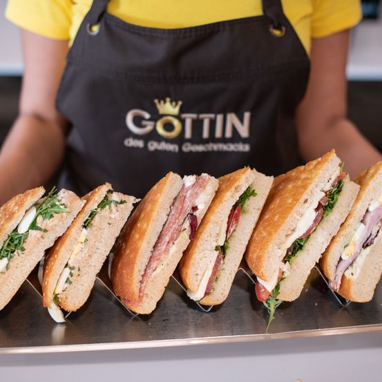 Eine Person in einer schwarzen Schürze mit Gottin-Logo präsentiert eine Reihe von Sandwiches auf einem silbernen Tablett. Die Sandwiches haben verschiedene Füllungen.