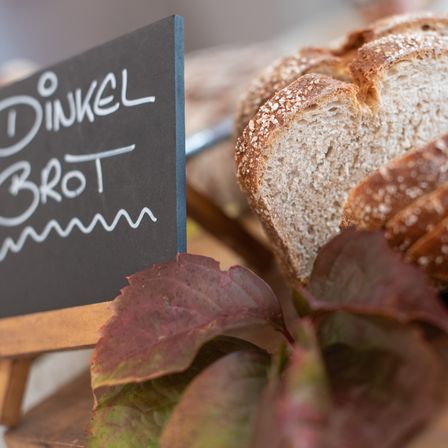 Ein Nahaufnahme eines Stücks rustikales Brot mit Körnern, begleitet von einem Schild mit der Aufschrift 'Dinkelbrot' und einigen roten Blättern.