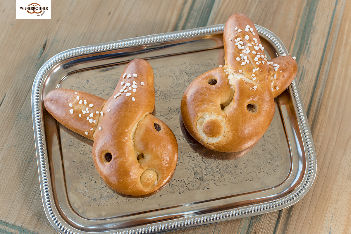 Zwei Osterbrote in Form von Häschen liegen auf einer silbernen Servierplatte mit einem dekorativen Muster. Sie sind mit weißem Zucker bestreut.