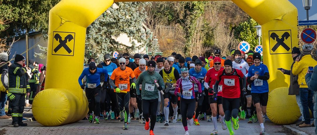 Marathonläufer in warmer Kleidung passieren einen gelben Bogen mit der Aufschrift 'Raiffeisenbank Baden'. Bäume und Schilder sind im Hintergrund zu sehen.
