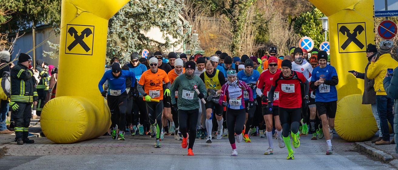 Eine Gruppe von Läufern mit nummerierten Startnummern nimmt an einem Marathon teil und passiert einen gelben Torbogen. Der Bogen trägt den Text 'Raiffeisenbank Baden' und ein Hammersymbol. Bäume und Straßenschilder sind im Hintergrund zu sehen.
