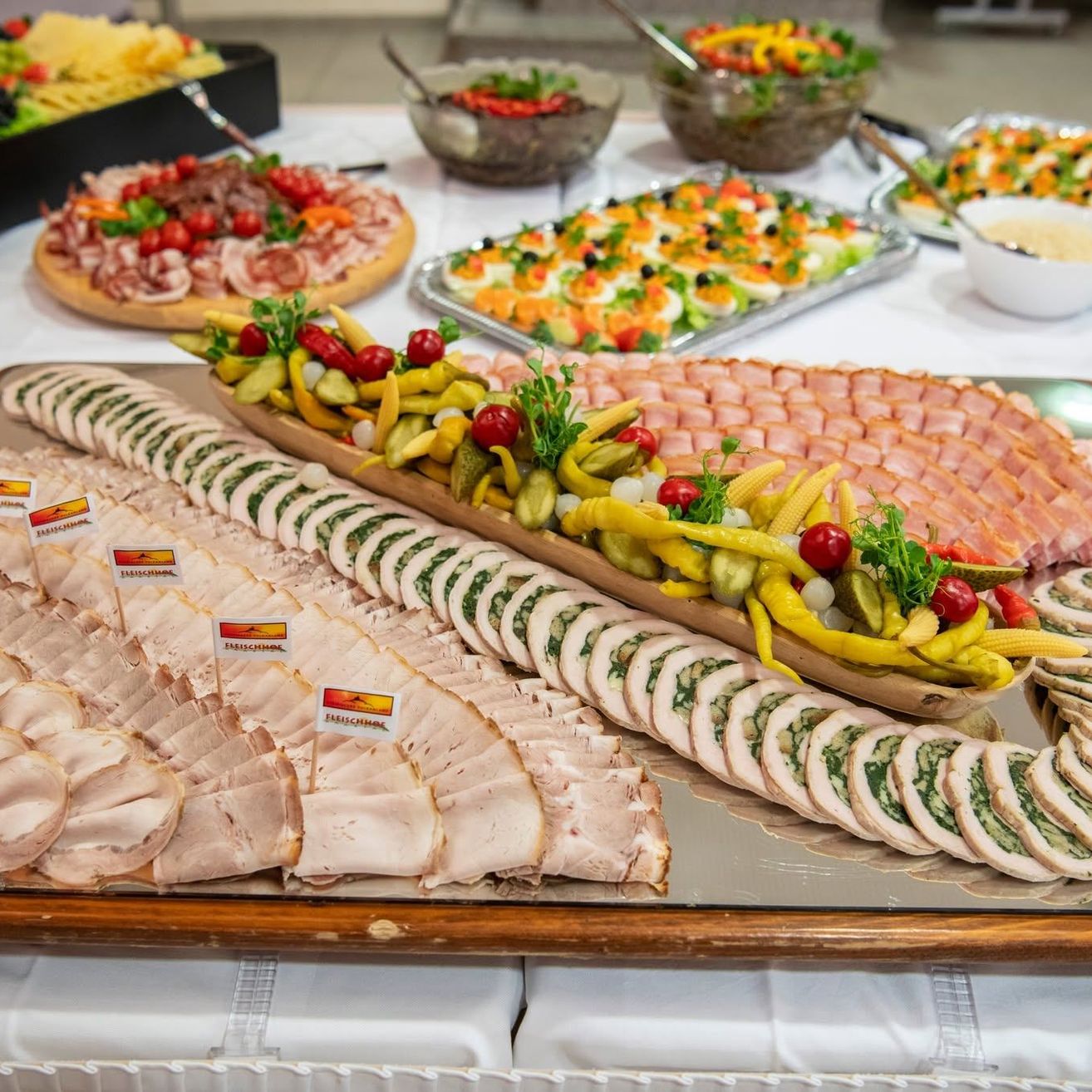 Ein Tisch mit verschiedenen Essensplatten, einschließlich geschnittener Fleischsorten, Gemüse und Garnierungen. Salatschüsseln und Dips sind ebenfalls vorhanden.