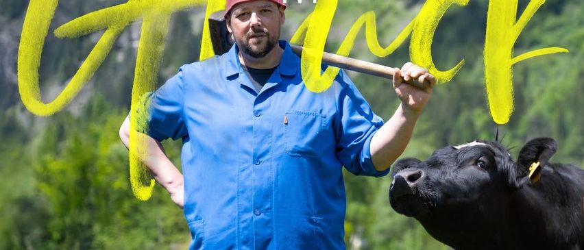 Ein Mann in einem blauen Hemd und einem roten Hut hält einen Holzstock in einem Feld mit Kühen und Bergen im Hintergrund. Der Text lautet 'HONS IM GRICK' und 'GEMEINDEFESTSAAL HOCHHUBER PEGGAU 14.11.2026 | 19:30 Uhr'.