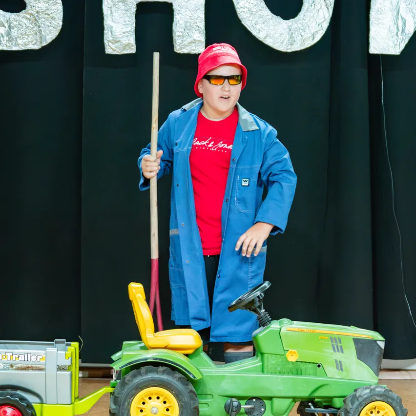Ein junger Junge in einem roten Shirt und einer blauen Jacke hält einen Stock und steht neben einem grünen Spielzeug-Traktor mit einem gelben Sitz.