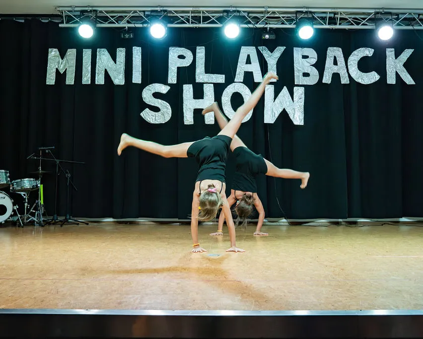 Zwei Frauen machen Handstände auf einer Bühne mit schwarzem Hintergrund und den Worten 'MINI PLAY BACK SHOW'. Links neben ihnen stehen Musikinstrumente.