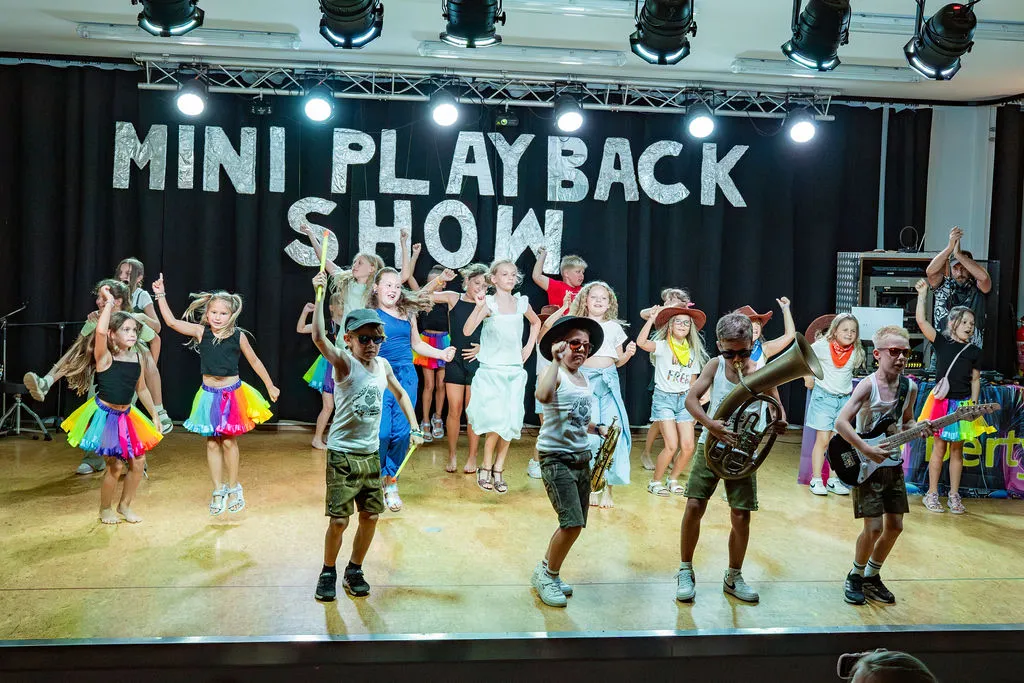 Auf einer Bühne führen Kinder eine Playback-Show auf. Sie tragen verschiedene Outfits und halten Musikinstrumente. Ein Banner steht 'Mini Playback Show'. Scheinwerfer beleuchten die Bühne.