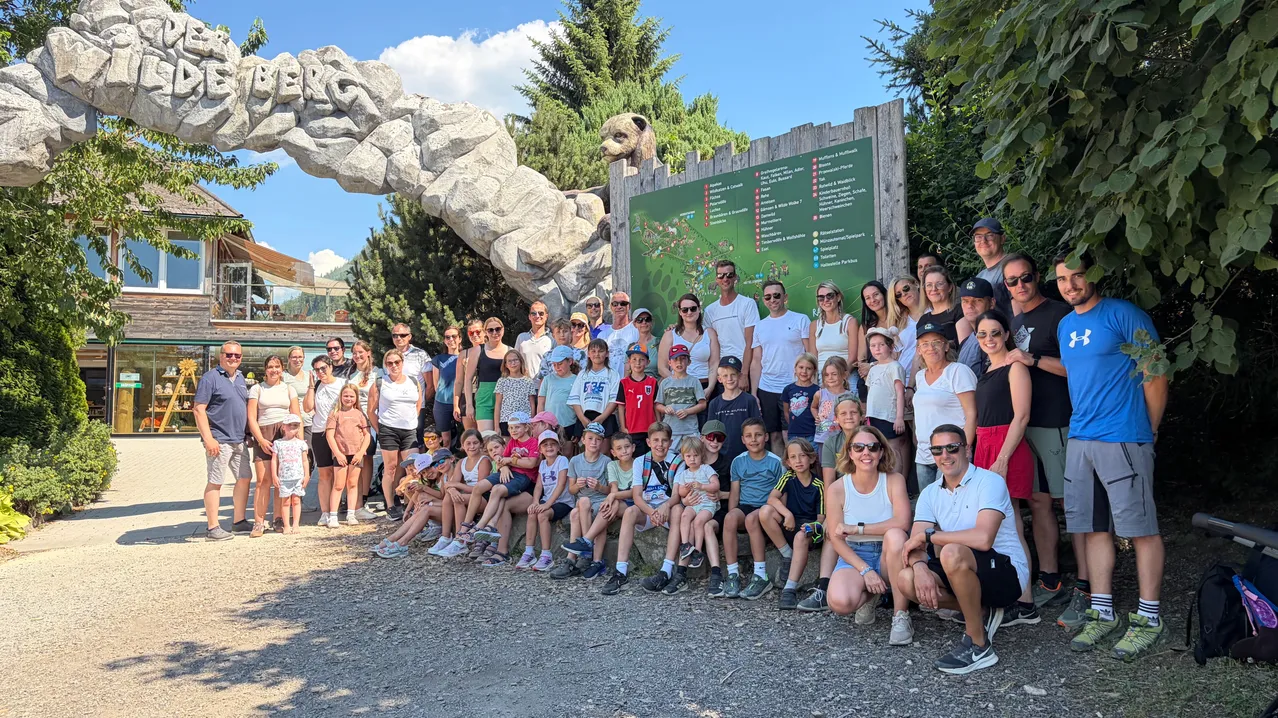 Eine große Gruppe von Menschen, einschließlich Erwachsener und Kindern, versammelt sich draußen für ein Foto vor einem Kartenschild. Das Schild ist von einer Felsstruktur umgeben.