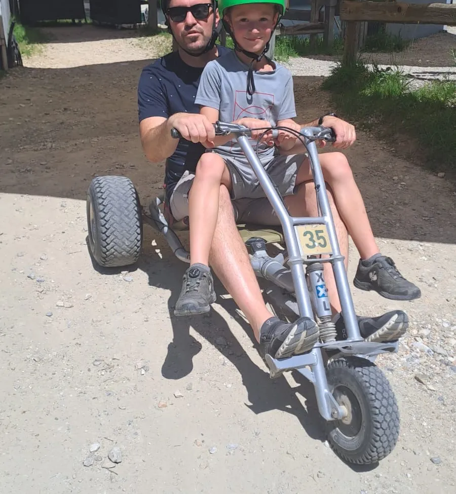 Ein Erwachsener und ein junger Junge fahren einen Go-Kart auf einer unbefestigten Straße, beide tragen Helme. Der Kart hat die Nummer 35 an der Front.