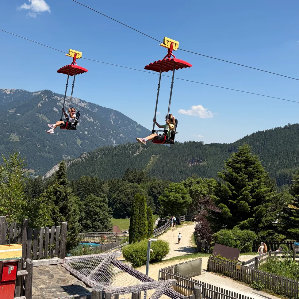 Zwei Personen fahren in einer roten Seilbahn mit gelben Oberteilen über einem Tal mit Bergen im Hintergrund.