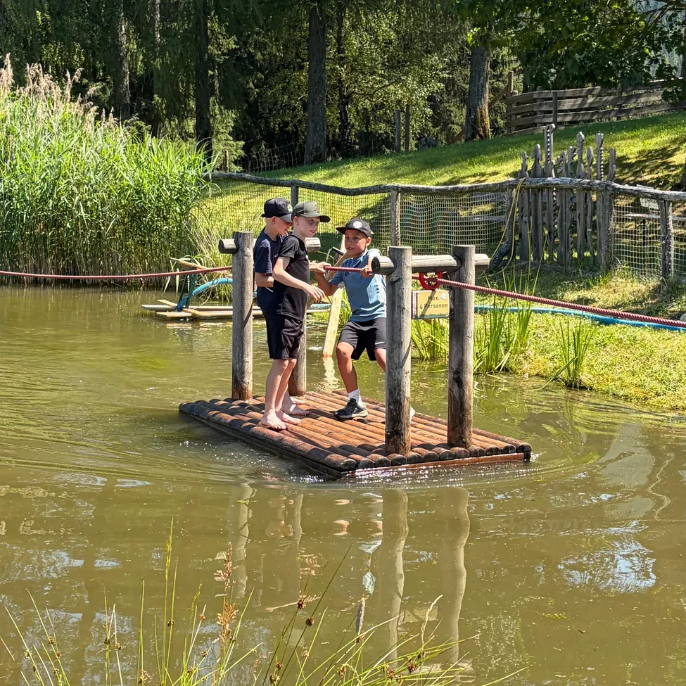 Drei Kinder spielen auf einer kleinen Holzbrücke über einem Teich. Sie sind barfuß. Hinter ihnen trennt ein Zaun den Rasen vom Wasser.