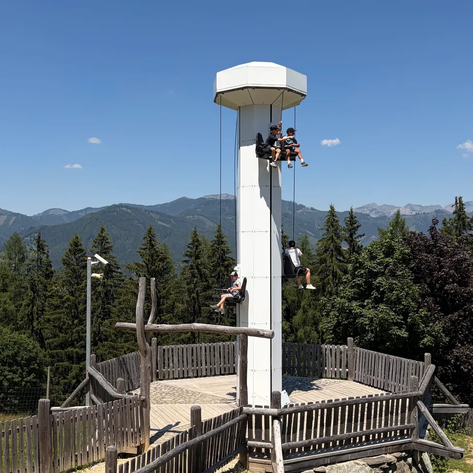 Drei Personen sitzen in einer Gondel auf der Spitze eines hohen Turms. Der Turm ist von einem Holzzaun mit einem Weg umgeben. Der Himmel ist klar mit ein paar Wolken. Im Hintergrund sind Berge und Bäume.