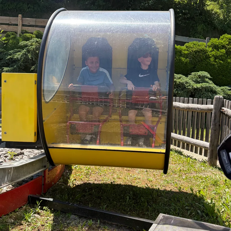 Zwei junge Jungen sitzen in einem durchsichtigen Fahrgeschäft in einem Vergnügungspark. Beide lächeln und halten das Lenkrad des Fahrgeschäfts.