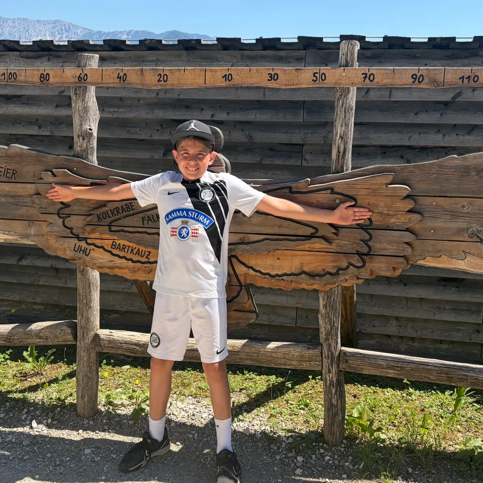 Ein junger Junge in einem Sportanzug steht unter einer Holzstruktur mit Flügeln und posiert für ein Foto. Hinter ihm ist ein Holzzaun mit Nummern und Namen zu sehen.