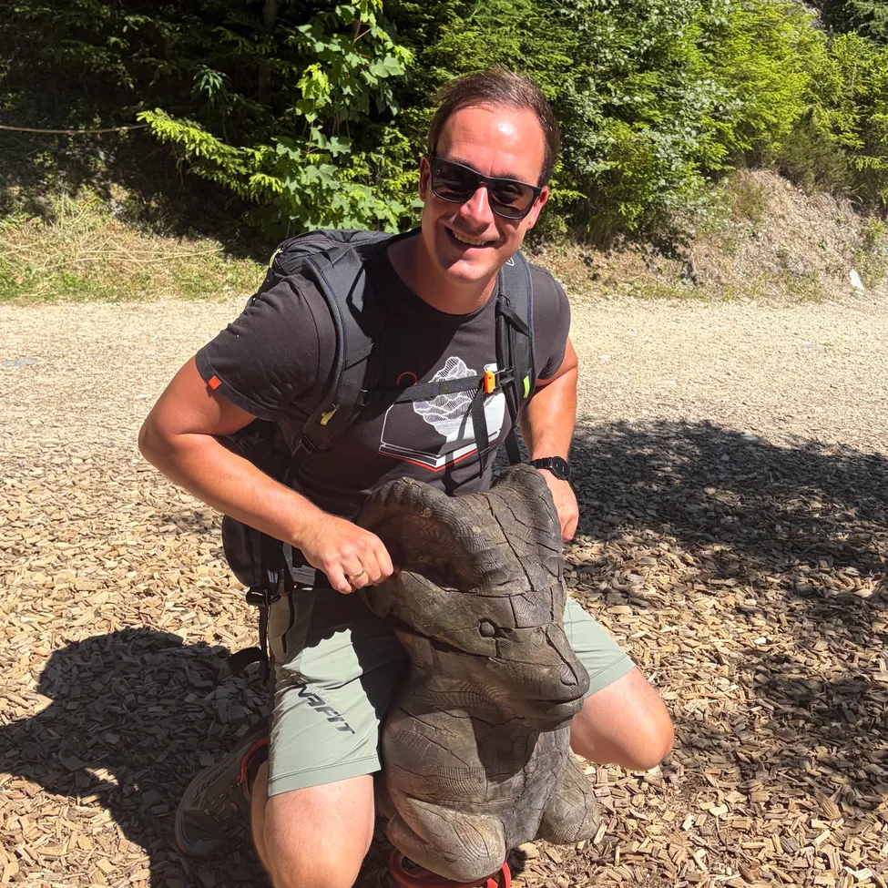 Ein Mann mit Rucksack sitzt auf einer geschnitzten Holzfigur eines Dinosauriers. Er lächelt und posiert für ein Foto an einem sonnigen Tag.