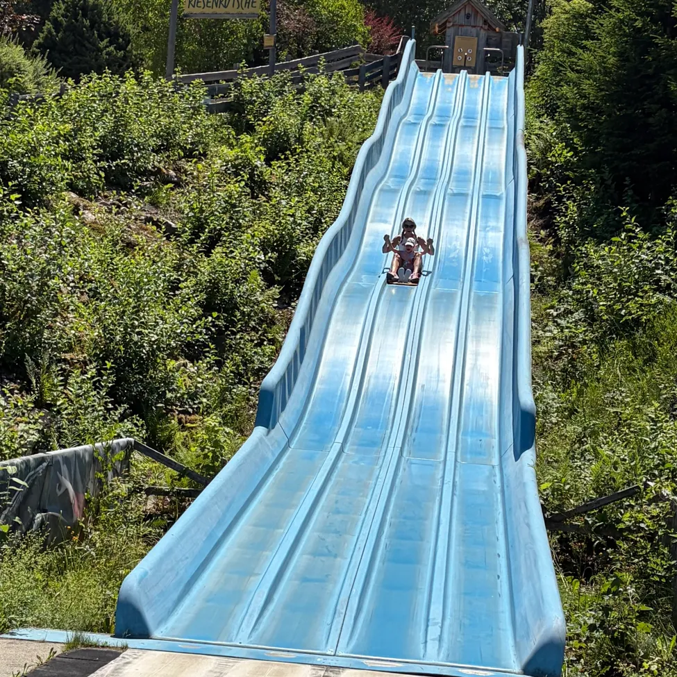 Eine blaue Wasserrutsche mit zwei Personen, die herunterrutschen. Umgeben von Grün und einem Holzzaun.