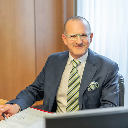 Ein Mann mit Brille sitzt lächelnd an einem Schreibtisch, hält einen Stift und ein Dokument. Er trägt einen grauen Anzug, ein weißes Hemd und eine gestreifte Krawatte. Hinter ihm steht eine Holzwand und ein Fenster mit Jalousien.