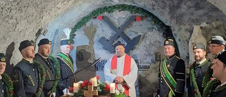 Ein Priester leitet eine Zeremonie in einer Höhle mit uniformierten Personen, Kerzen und Blumen auf einem Tisch, wahrscheinlich ein Weihnachtsereignis.