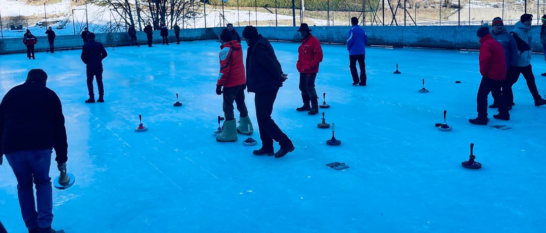 Eine Gruppe von Menschen in Winterkleidung spielt auf einer Eisbahn ein Curlingspiel. Die Bahn ist von einem Zaun umgeben, mit Bäumen und Häusern im Hintergrund.