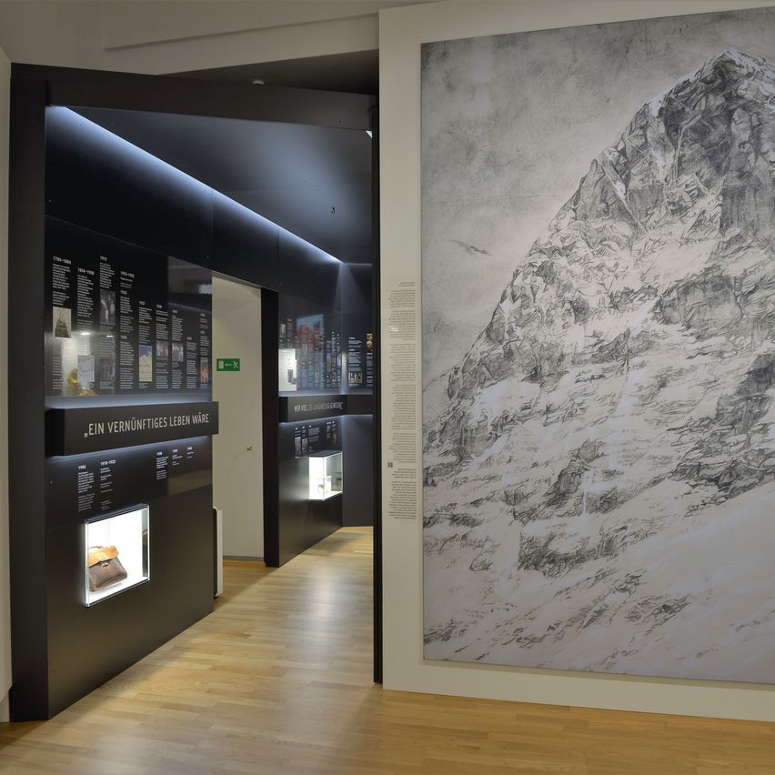 Ein Innenraum mit einer großen Zeichnung eines Berges an der Wand, einem Holzboden und einer schwarzen Vitrine mit einem Gegenstand darin.