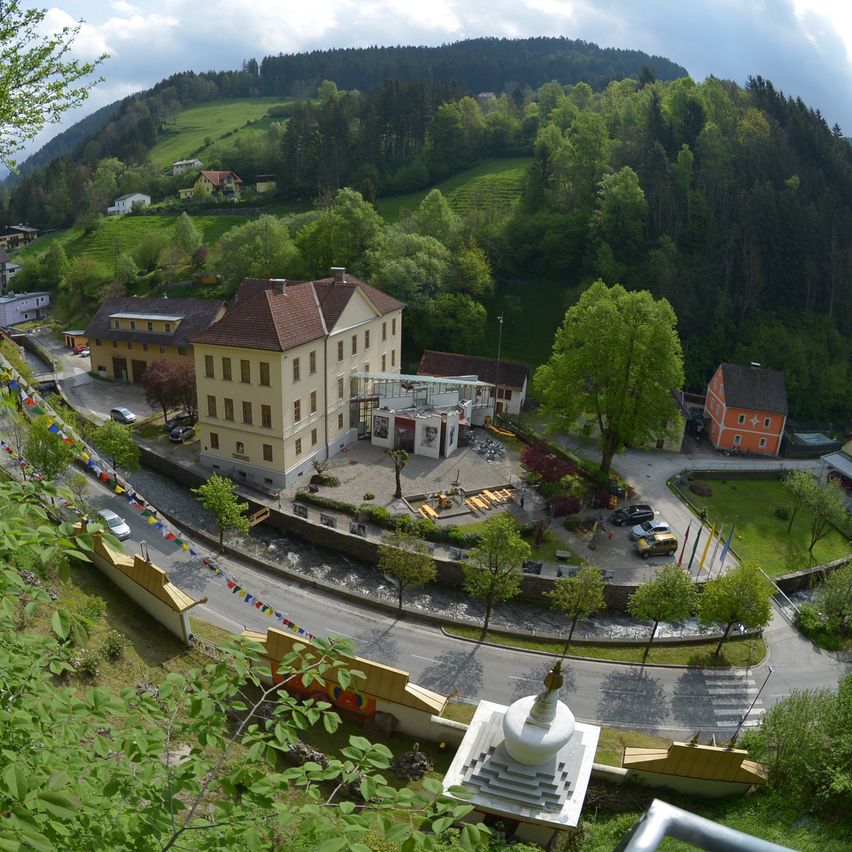 Luftaufnahme eines Dorfes mit einem großen Gebäude und kleineren Häusern in einer bergigen, bewaldeten Gegend. Die Straße hat geparkte Autos und Flaggen.