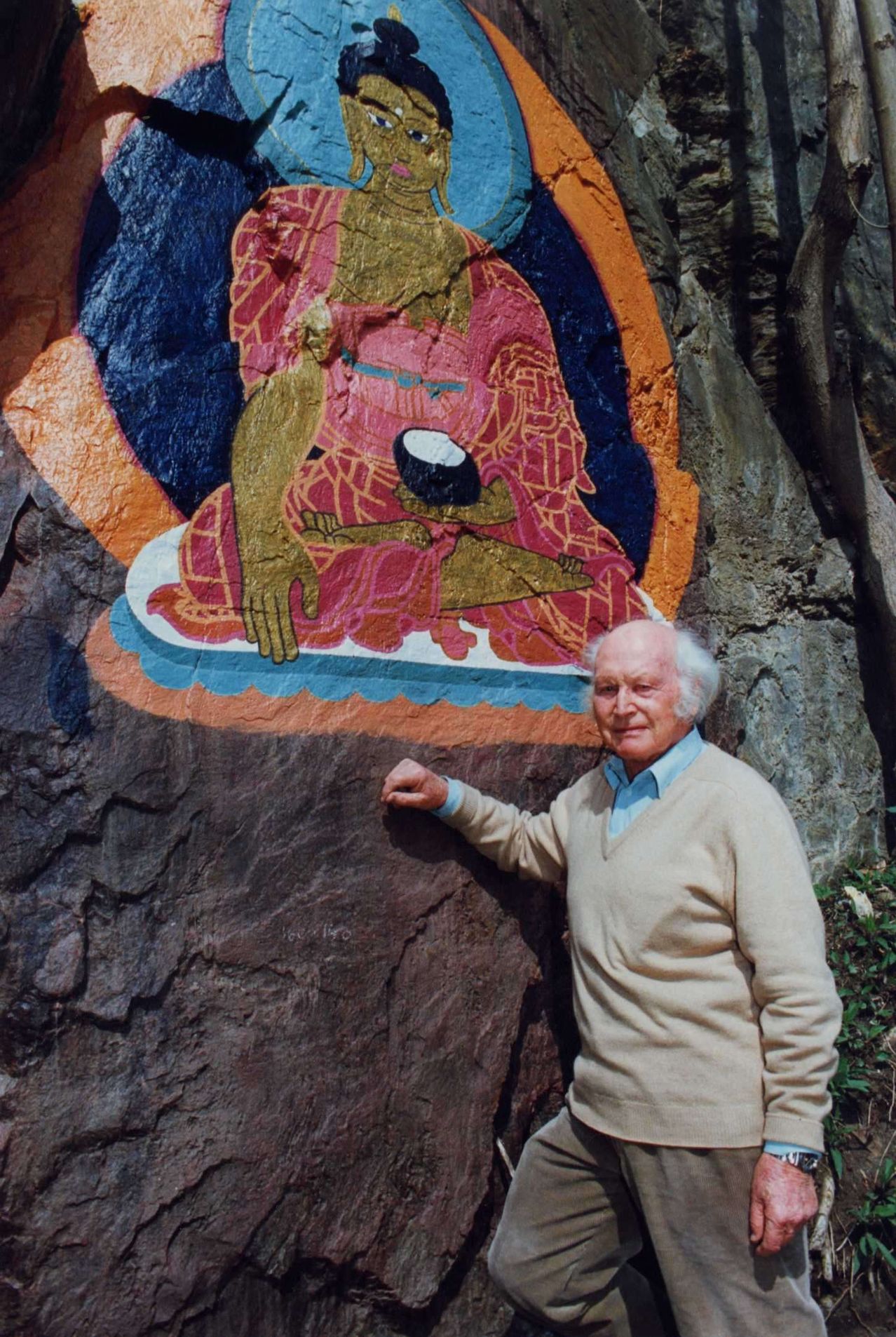Ein älterer Mann steht neben einem Felsbrocken mit einer farbenfrohen buddhistischen Figur, die darauf gemalt ist. Er trägt einen beigen Pullover und ein blaues Hemd.